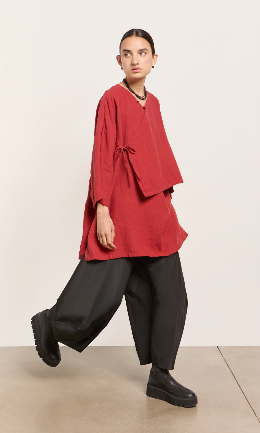 Plümo  Model wears the Leila tunic in poppy red, paired with black pants and platform shoes, walking indoors on a beige floor.