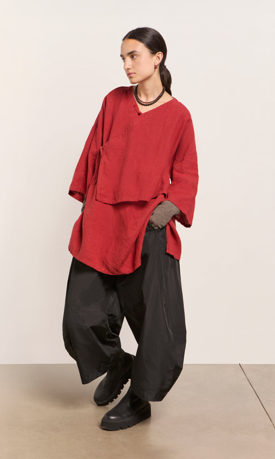 Plümo  Person in a poppy red Leila tunic, black wide pants, and boots stands before a plain light backdrop, gazing aside.