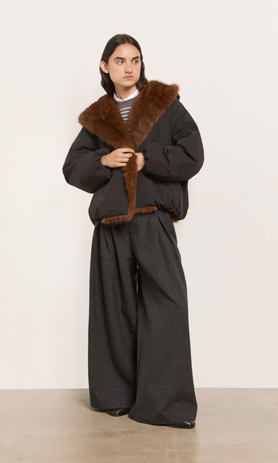 Plümo  Model in an acorn brown Lenz Parka with oversized hood and wide-leg gray pants stands against a plain background.