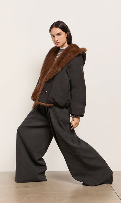 Plümo  Model in the acorn brown Lenz Parka with an oversized hood and gray wide-leg pants poses against a light backdrop.