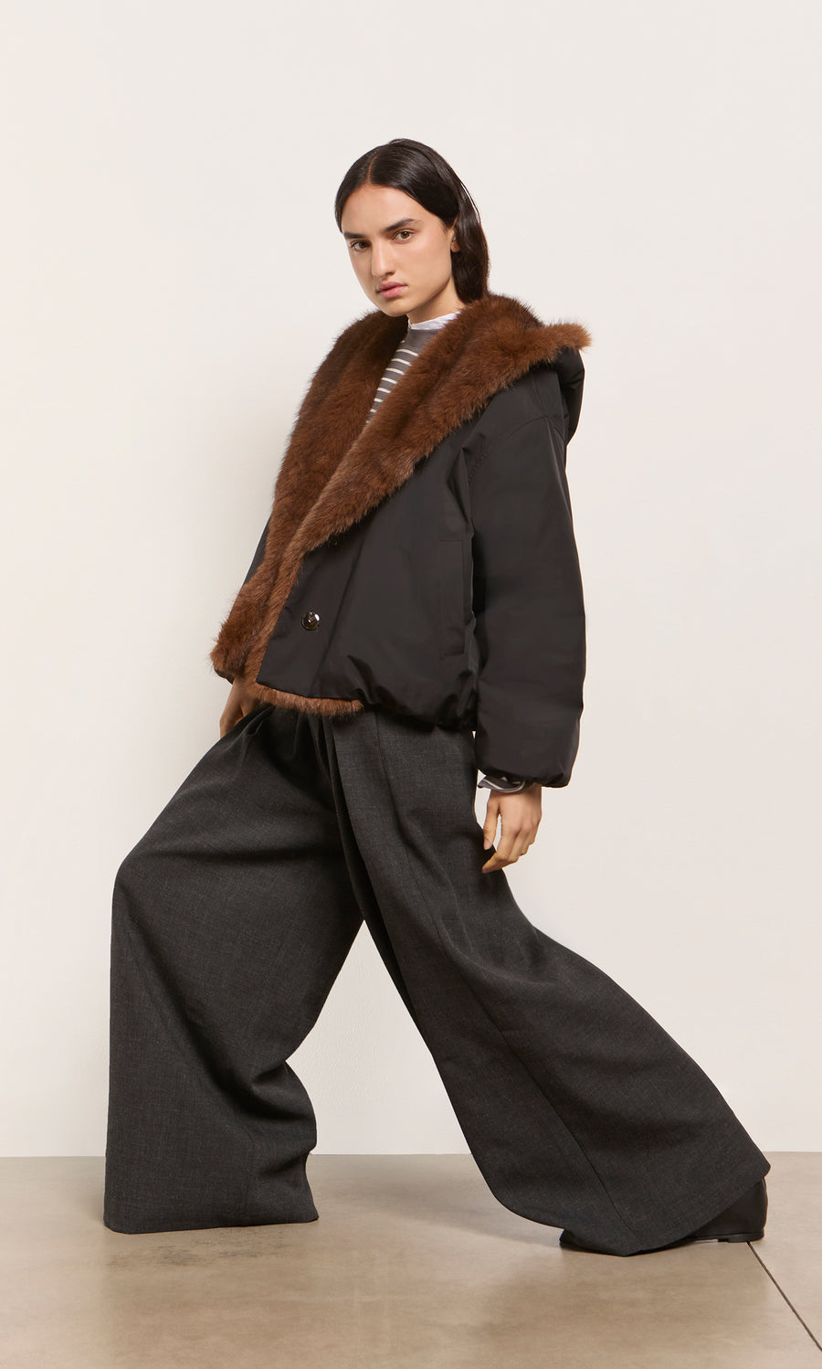 Plümo  Model in the acorn brown Lenz Parka with an oversized hood and gray wide-leg pants poses against a light backdrop.