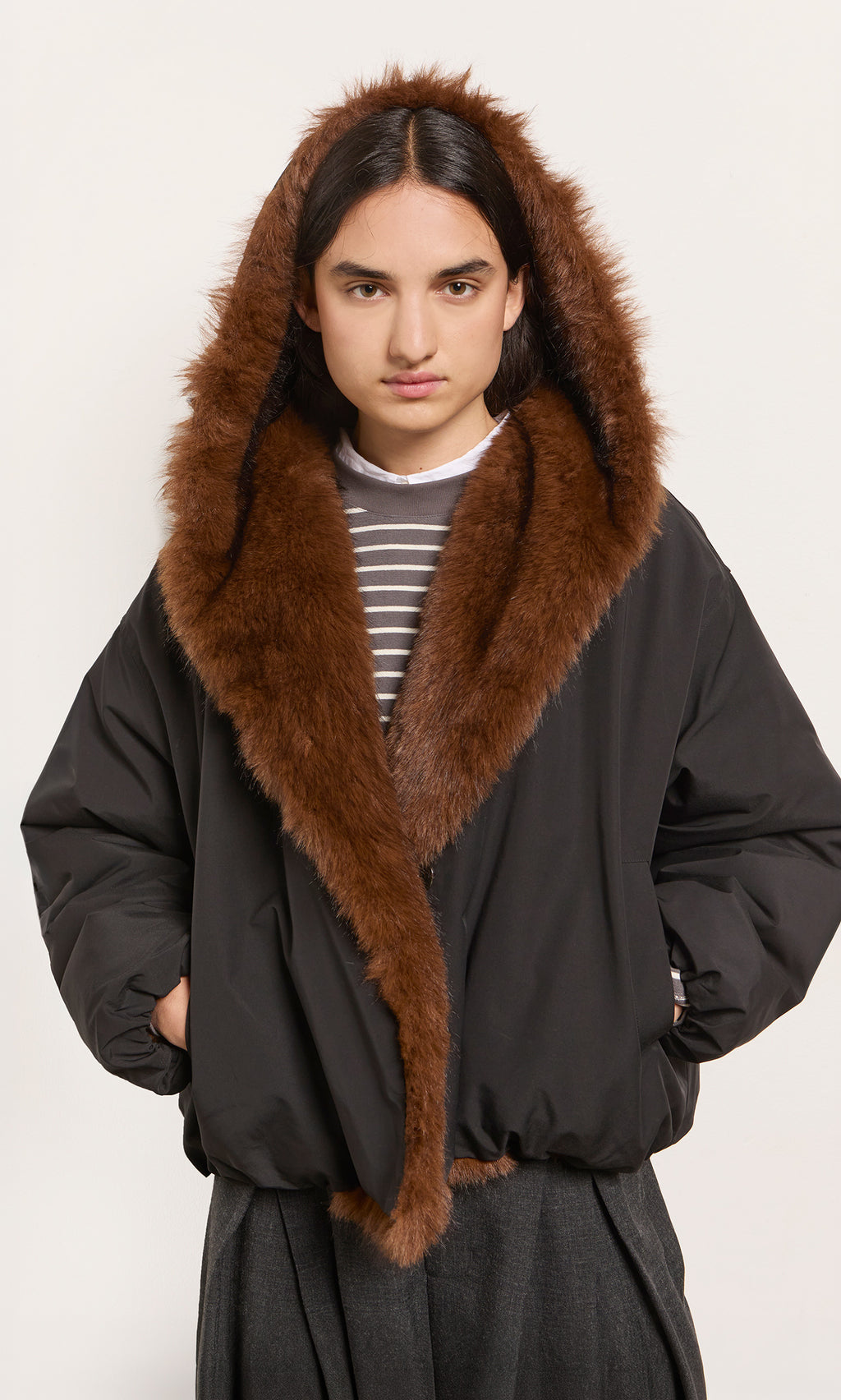 Plümo  Model wears the Lenz Parka in acorn brown, featuring faux fur and an oversized hood, hands in pockets.