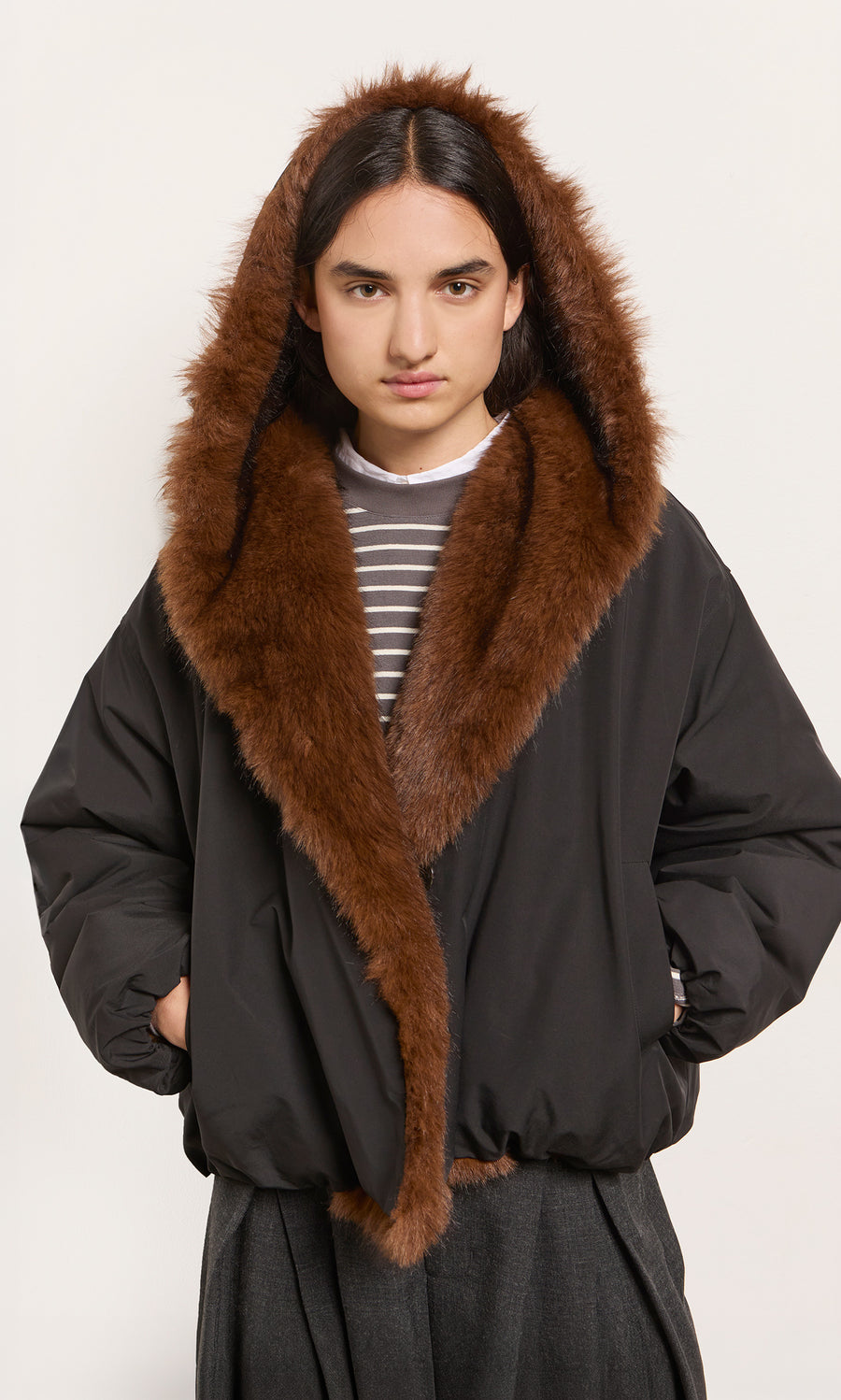 Plümo  Model wears the Lenz Parka in acorn brown, featuring faux fur and an oversized hood, hands in pockets.