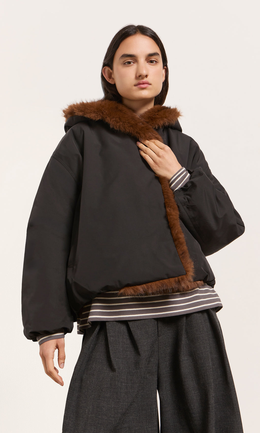 Plümo  A person in the acorn brown Lenz Parka, wide gray pants, and a striped shirt poses against a plain backdrop.
