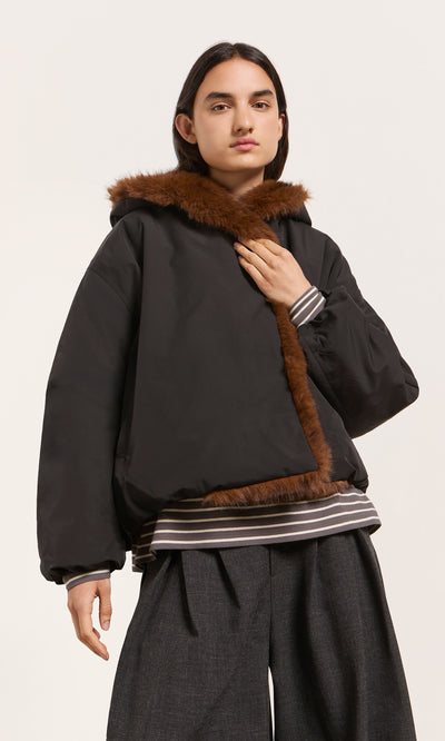 Plümo  A person in the acorn brown Lenz Parka, wide gray pants, and a striped shirt poses against a plain backdrop.