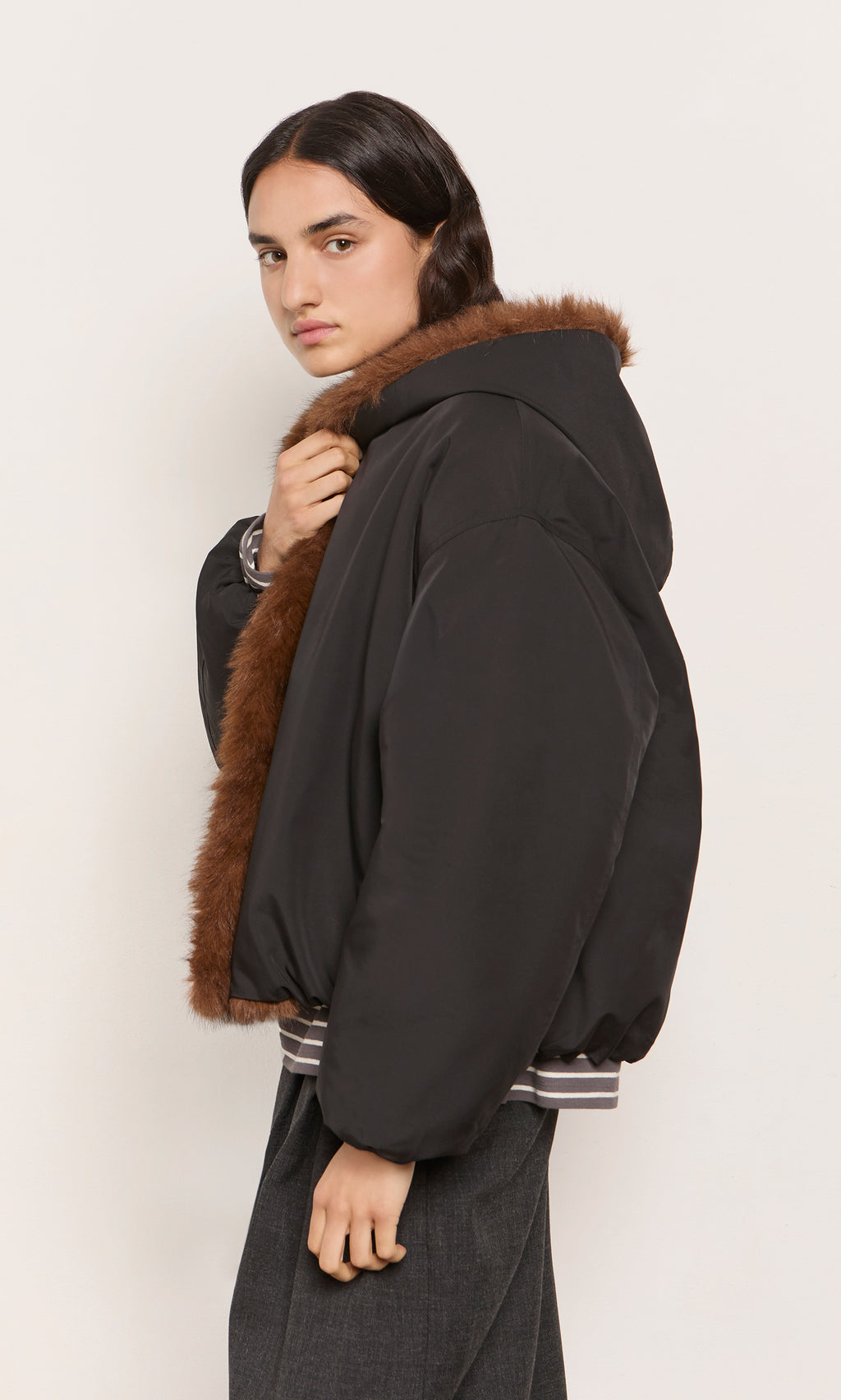 Plümo  Model in Lenz Parka, acorn brown with oversized hood, looks over their shoulder at the camera.