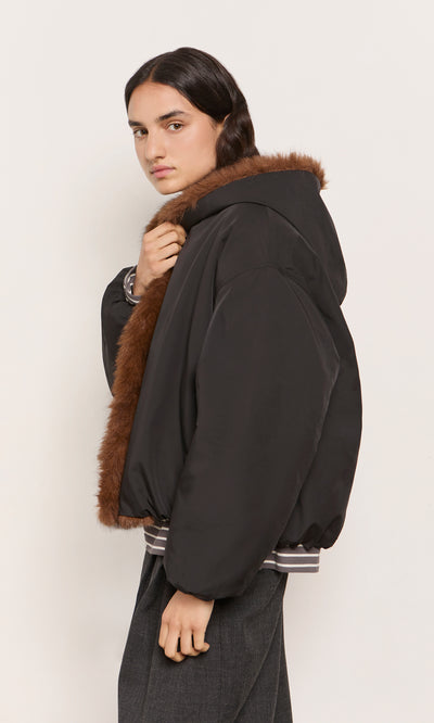 Plümo  Model in Lenz Parka, acorn brown with oversized hood, looks over their shoulder at the camera.