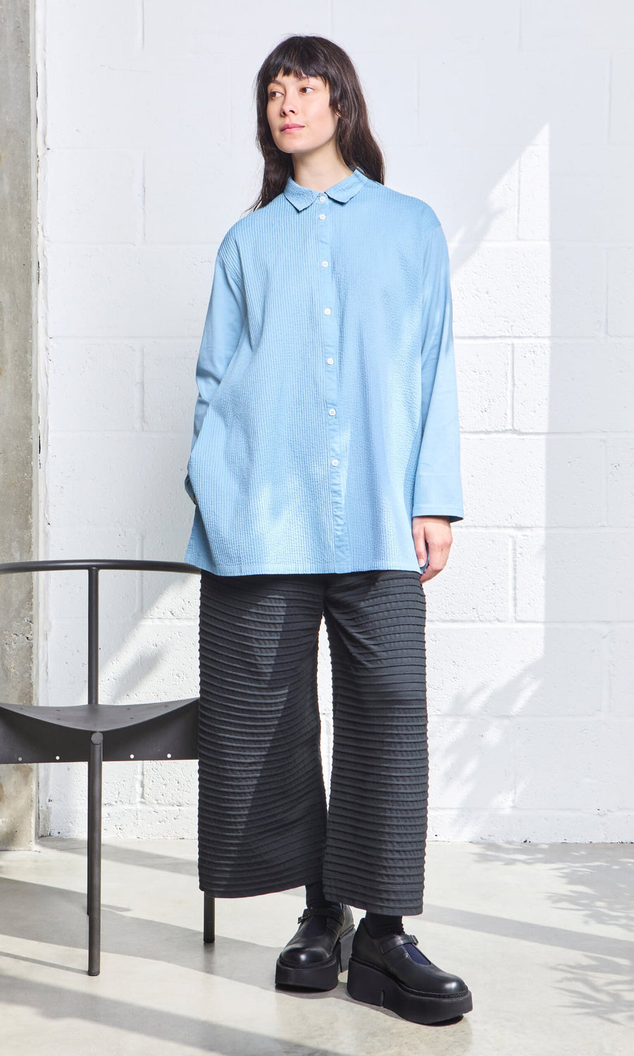 Plümo- Woman in the Leung Shirt and black wide-leg pants, standing indoors beside a black chair.