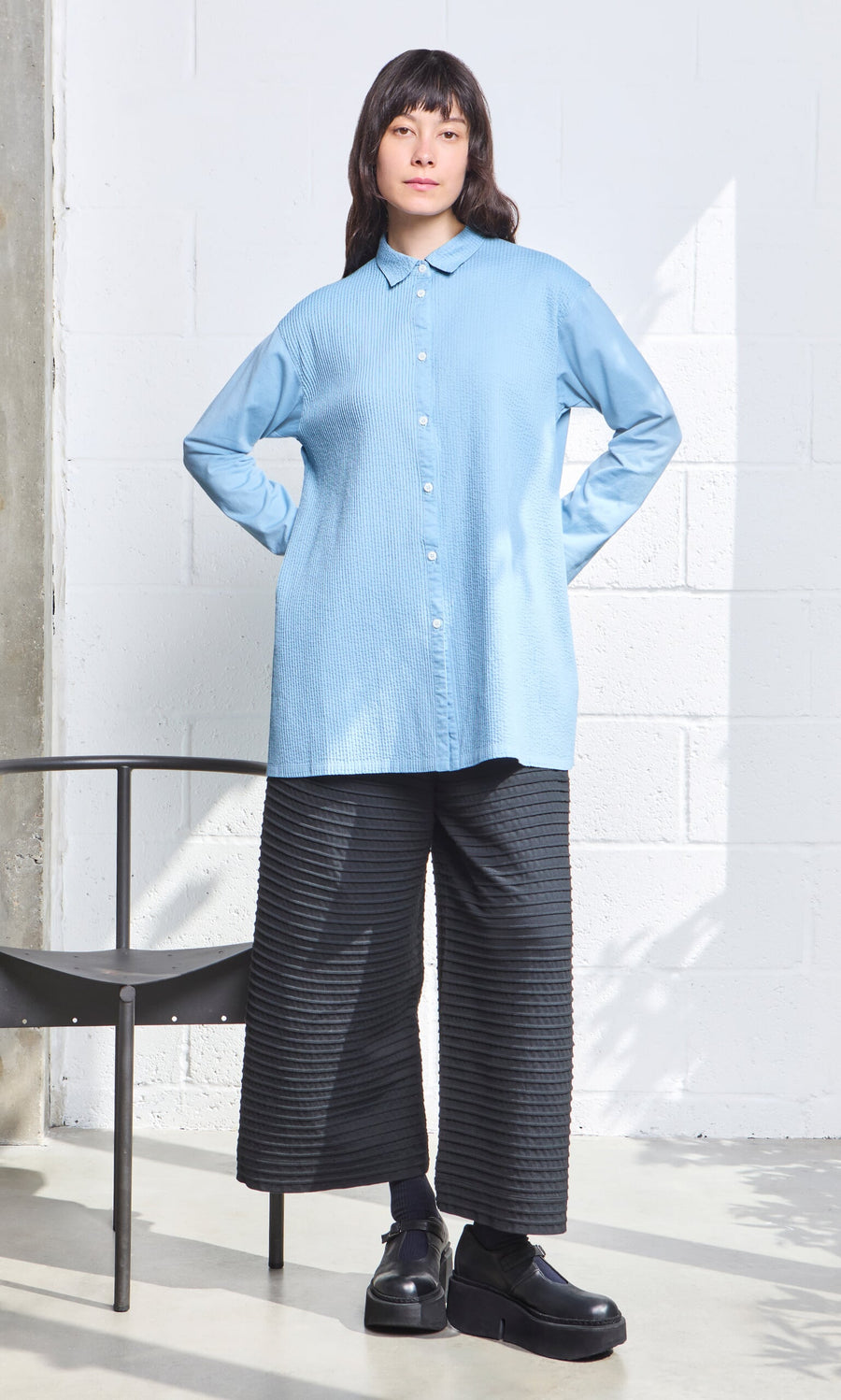 Plümo- Woman in a Yacco Maricard Leung Shirt and black wide-leg pants stands indoors, hands on hips by a metal chair.