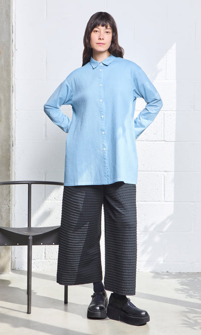 Plümo- Woman in a light blue button-up shirt and Koji Pants stands indoors next to a black chair.
