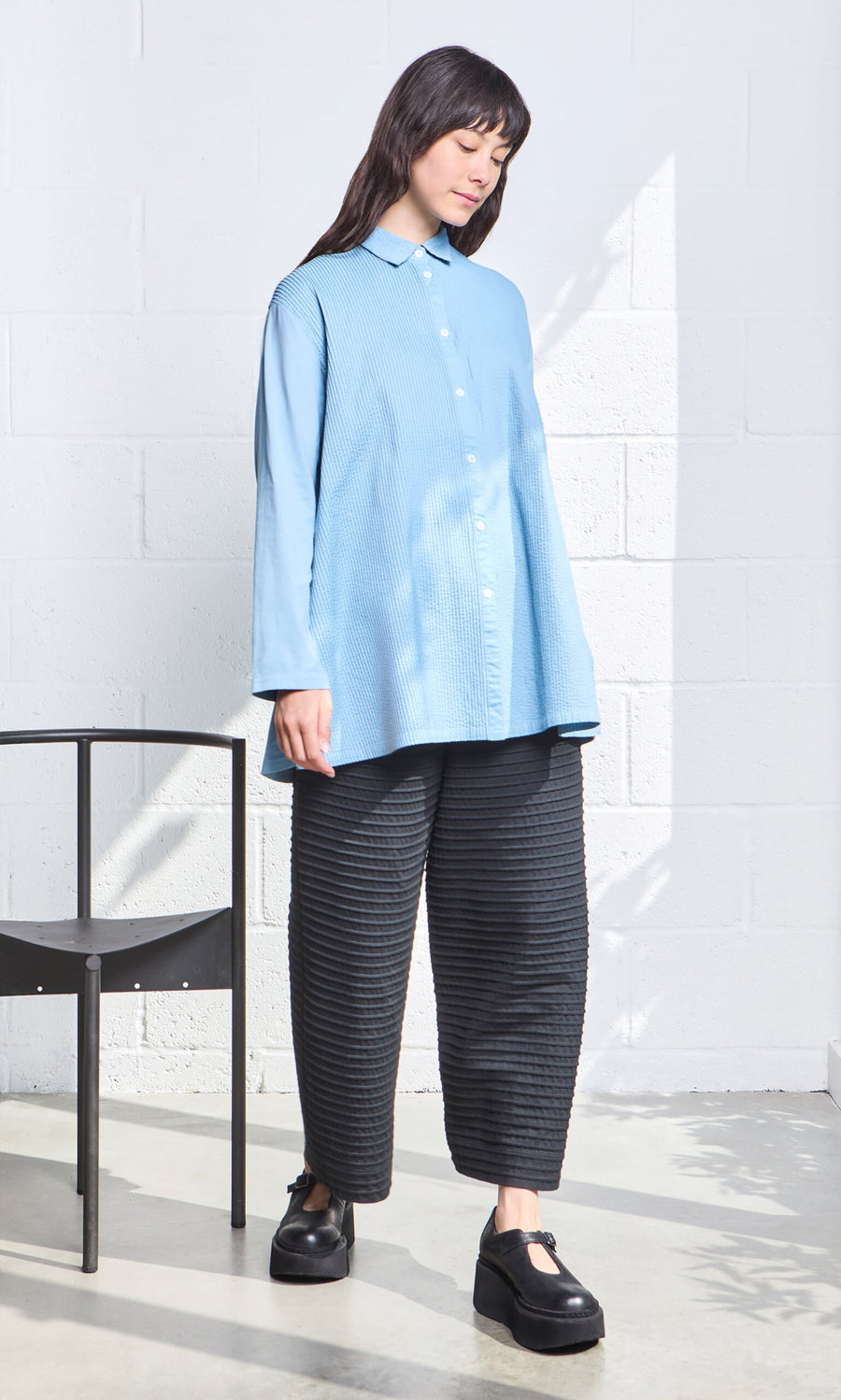 Plümo- Woman in a Leung Shirt and black pants stands by a black chair in a bright room.