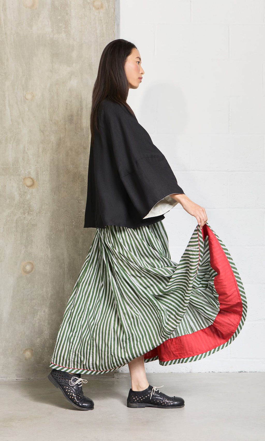 Plümo- A woman in a black jacket and the Lilia Skirt walks by a concrete and white wall background.