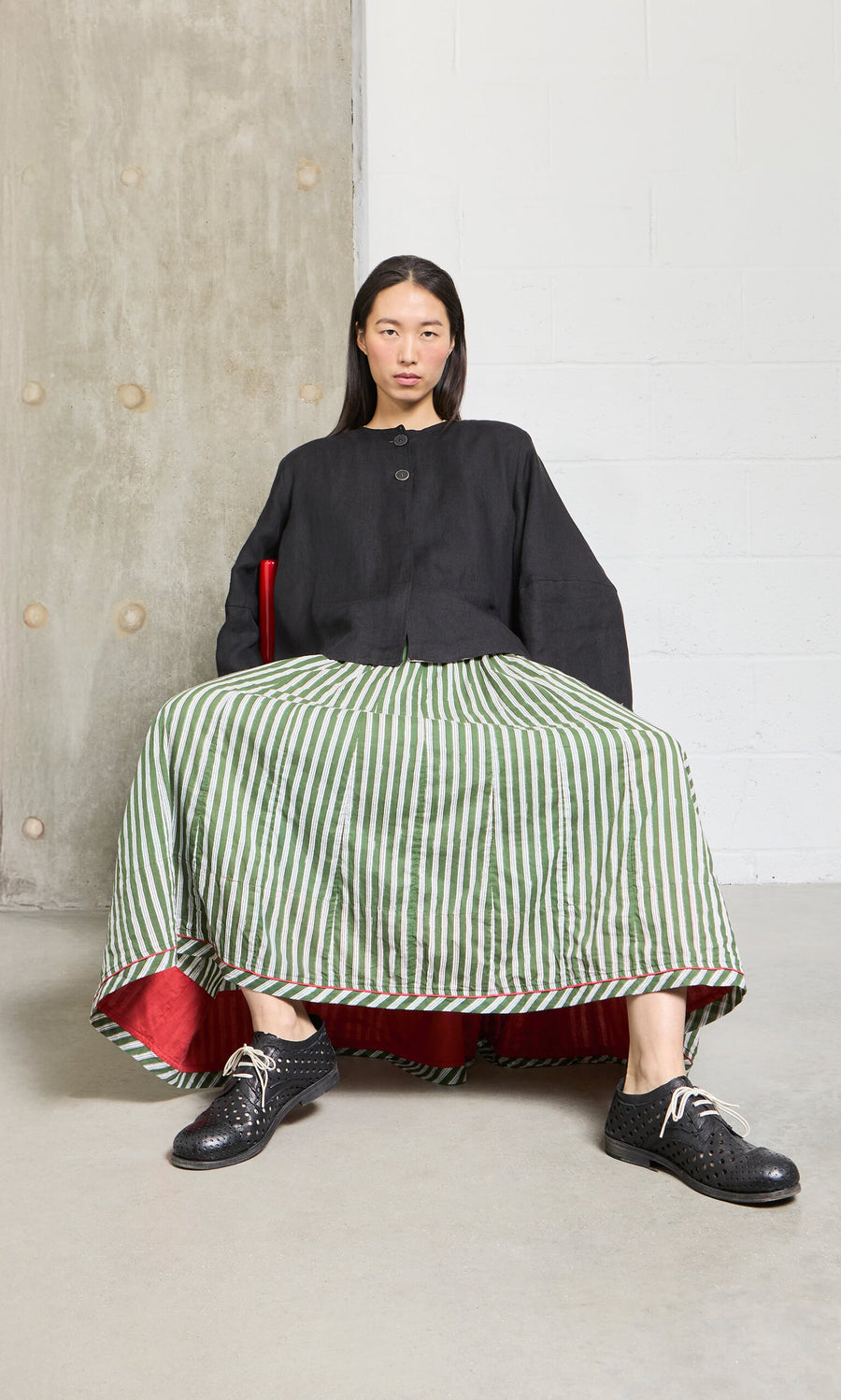 Plümo- Person in a black top and green Lilia Skirt with elastic waistband sits on a red chair, looking at the camera.