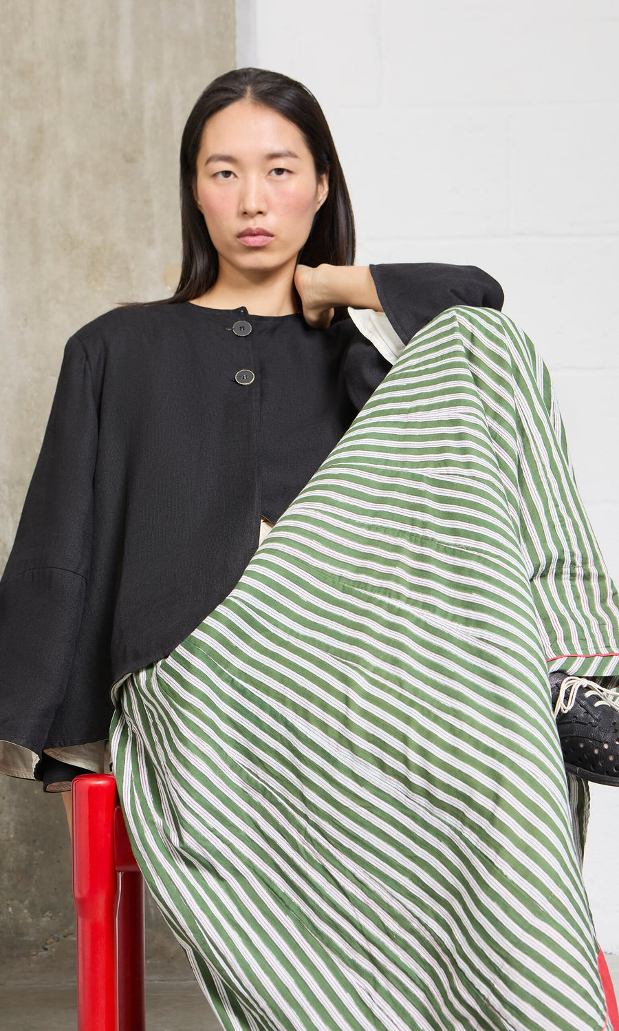 Plümo- Woman in black top and Lilia Skirt with green-and-white stripes sits on red chair, looking at the camera.