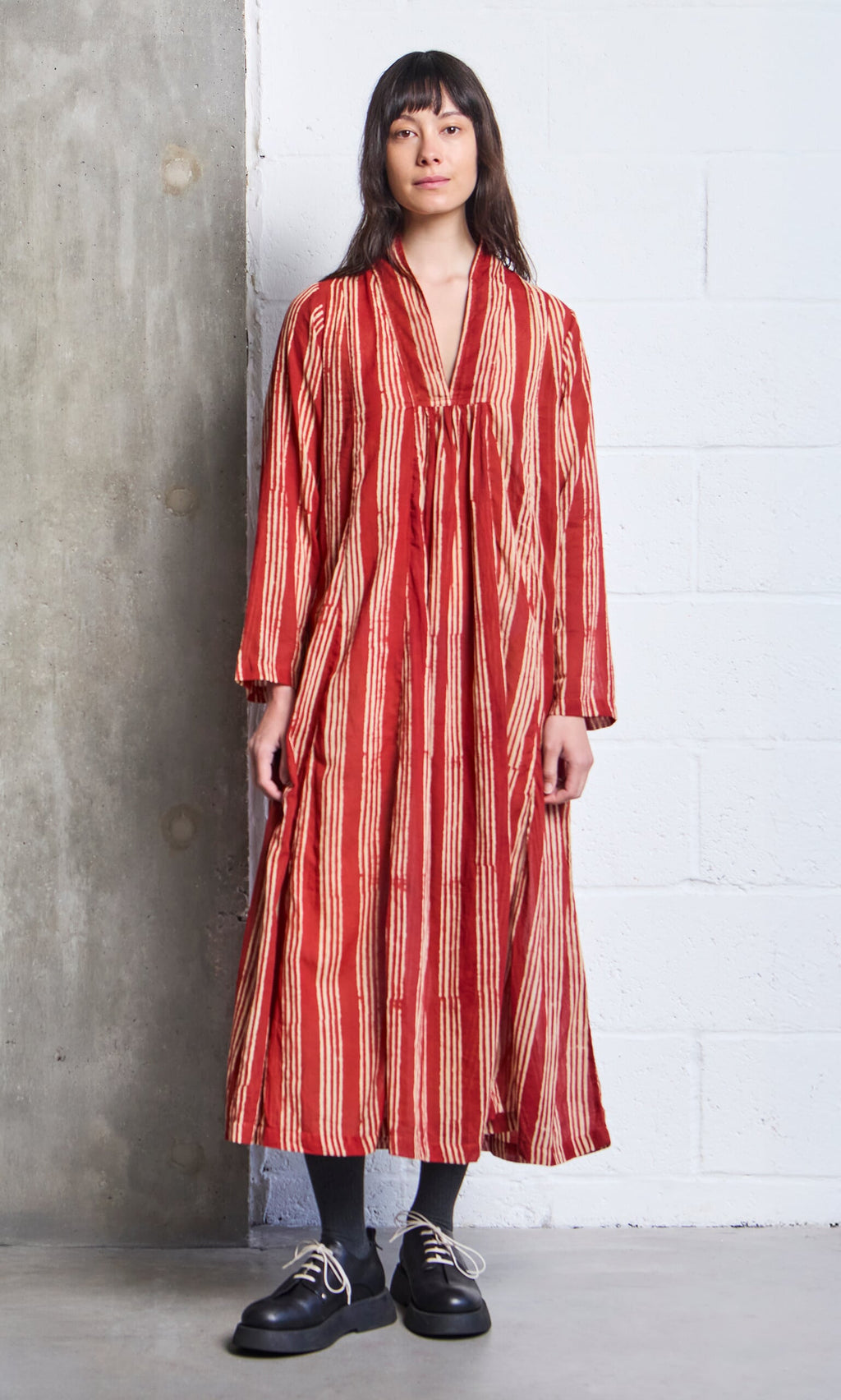 Plümo- Woman wearing the Lina Stripe Dress, a long, loose block-printed A-line with vertical cream stripes, standing indoors.