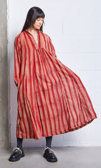 Plümo- Woman in a Lina Stripe Dress—an A-line, long red block-printed dress with white stripes—stands by a wall.