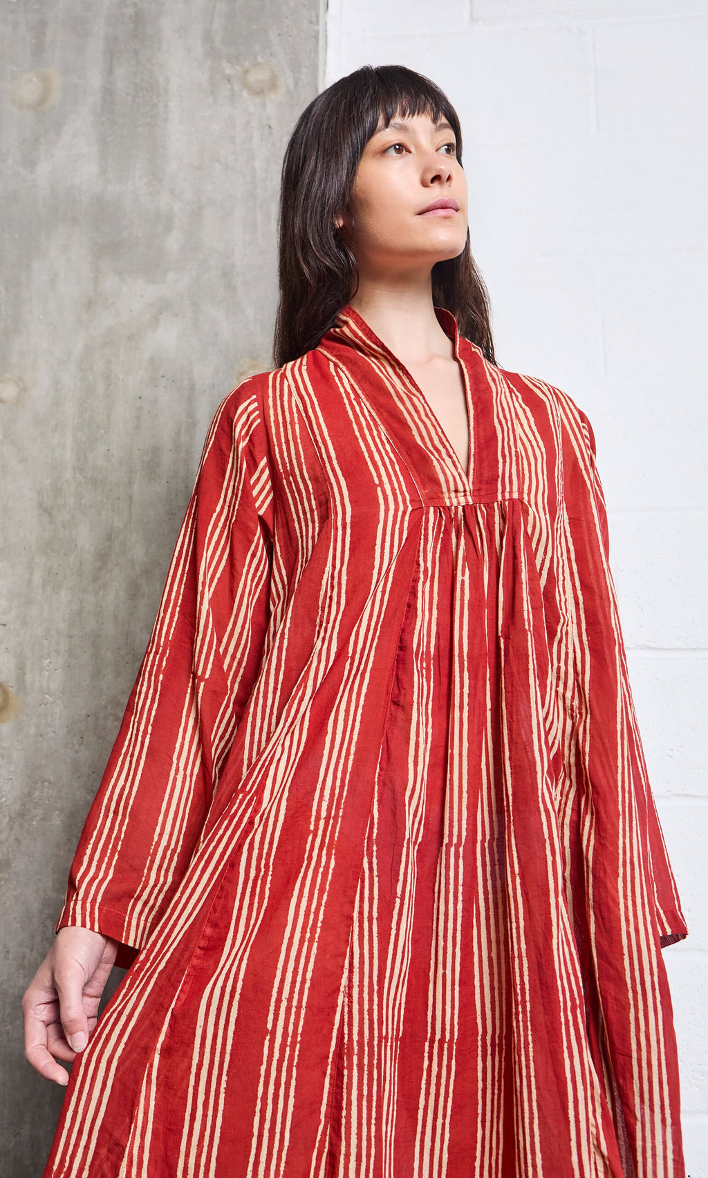 Plümo- Woman in a loose-fitting Lina Stripe Dress stands against a textured wall, looking upwards.