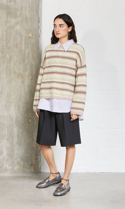 Plümo- Woman in a Lint Cardigan over a collared shirt, black knee-length shorts, and metallic shoes standing indoors.