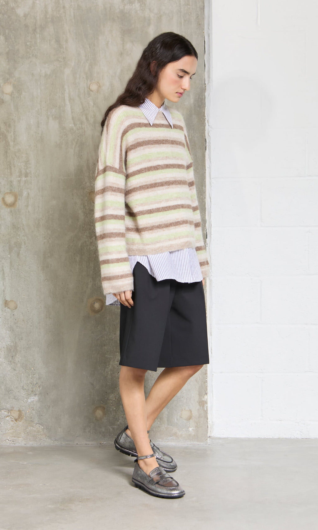 Plümo- A person in a Lint Cardigan, collared shirt, black shorts, and metallic shoes stands indoors by a wall.