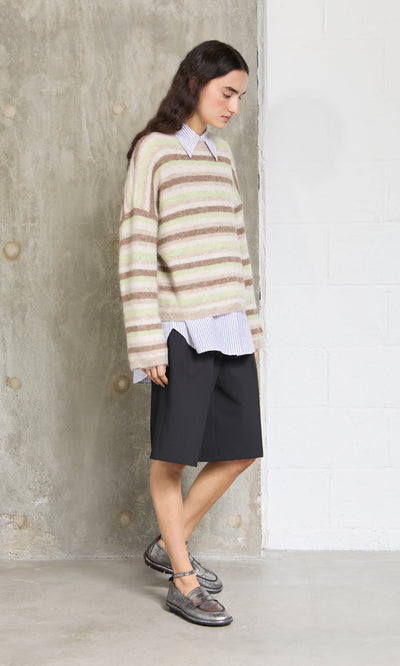 Plümo- A person in a Lint Cardigan, collared shirt, black shorts, and metallic shoes stands indoors by a wall.