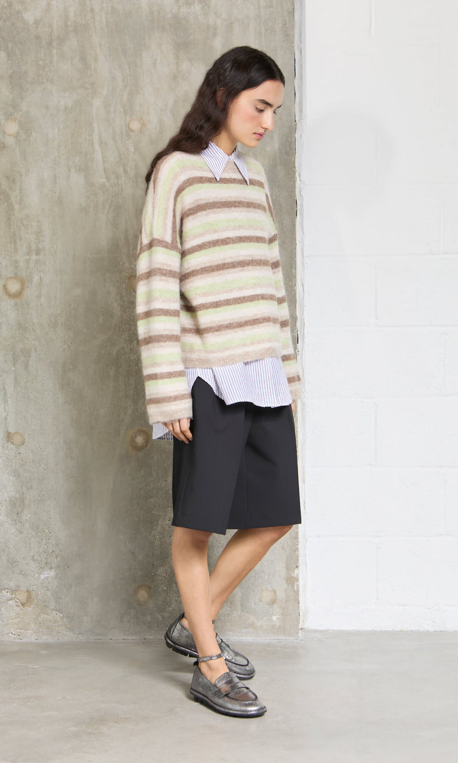 Plümo- A person in a Lint Cardigan, collared shirt, black shorts, and metallic shoes stands indoors by a wall.