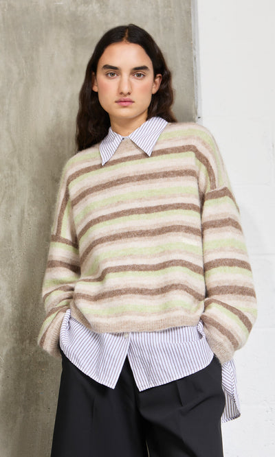 Plümo- Someone in the Lint Cardigan over a collared shirt and black pants stands by a white and concrete wall.