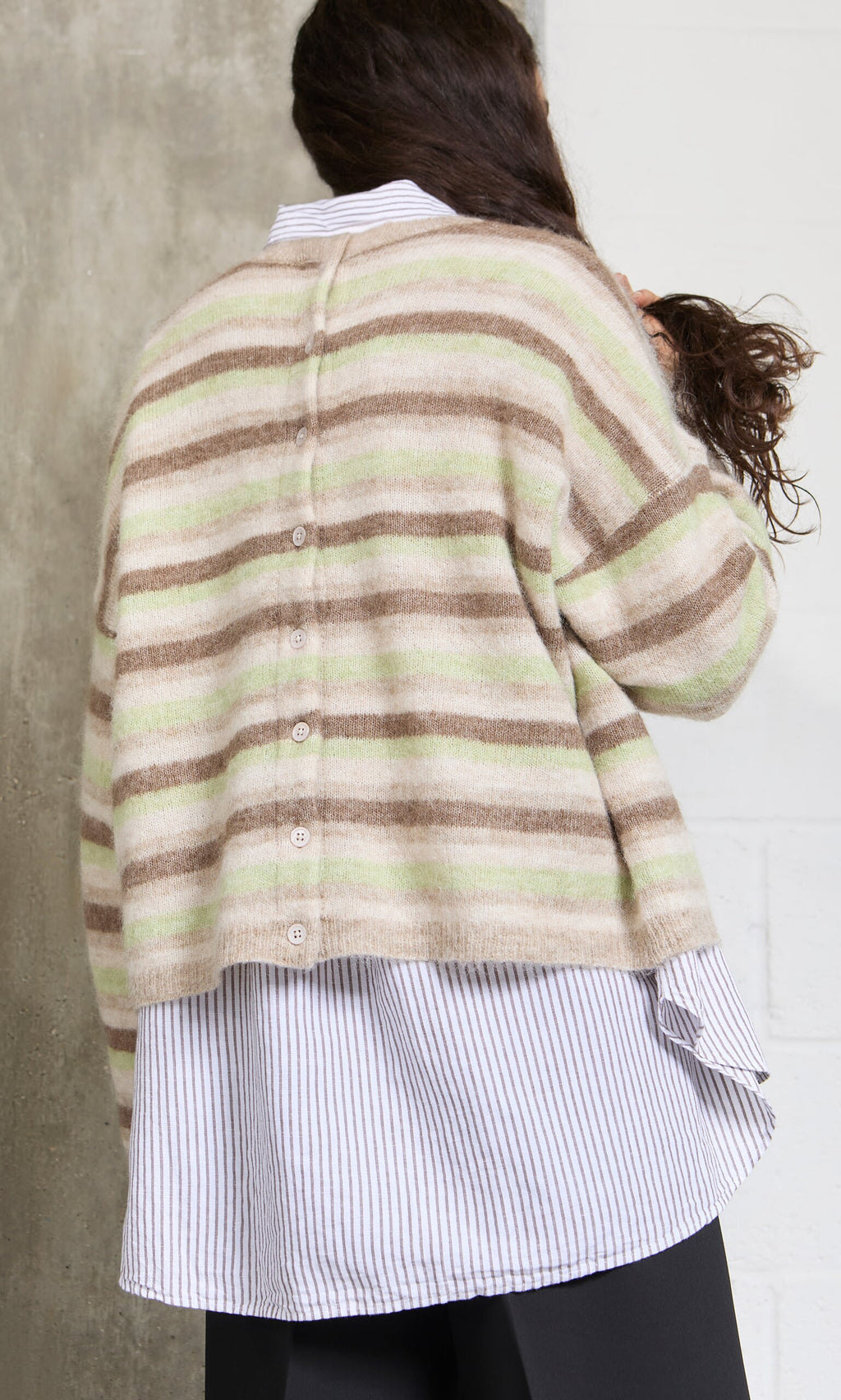 Plümo- A woman faces away, wearing the Lint Cardigan over a striped shirt and dark pants.