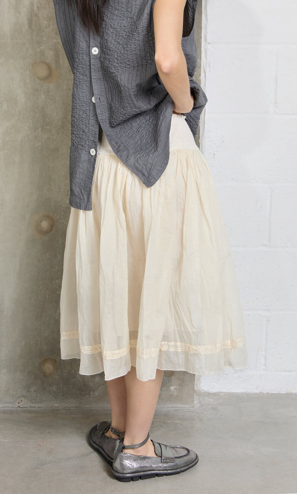 Plümo- A woman in a gray top and Lou Organza Skirt stands indoors, facing away, wearing metallic flat shoes.