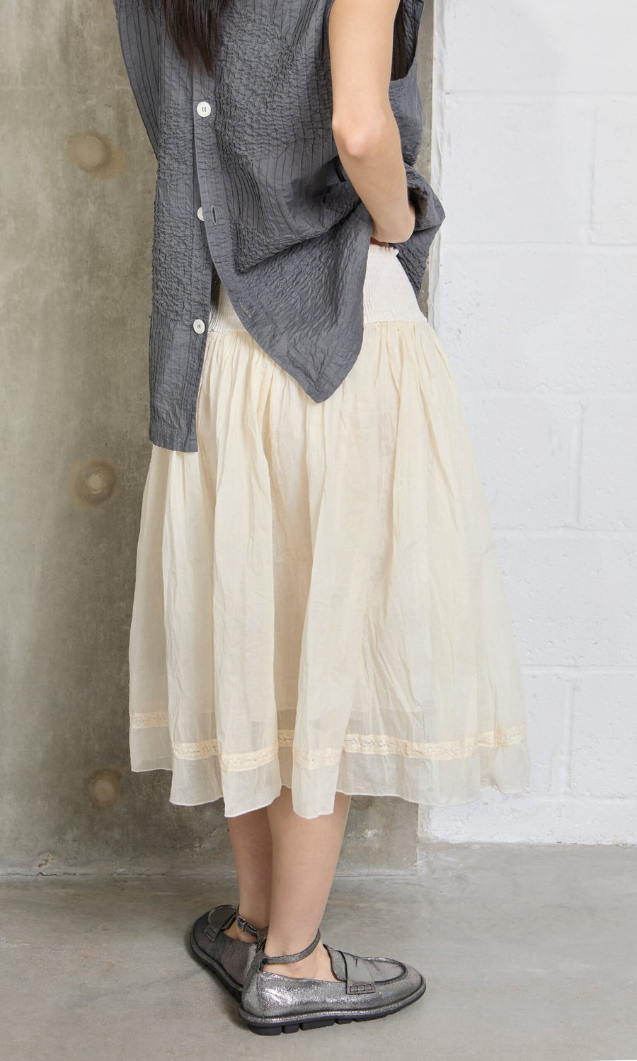 Plümo- A woman in a gray top and Lou Organza Skirt stands indoors, facing away, wearing metallic flat shoes.