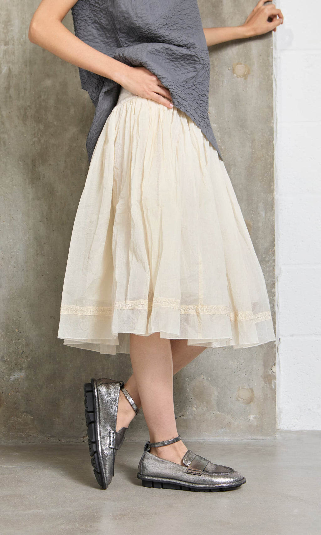 Plümo- Person in a grey top, Lou Organza Skirt in cream, and metallic shoes stands by a concrete wall.