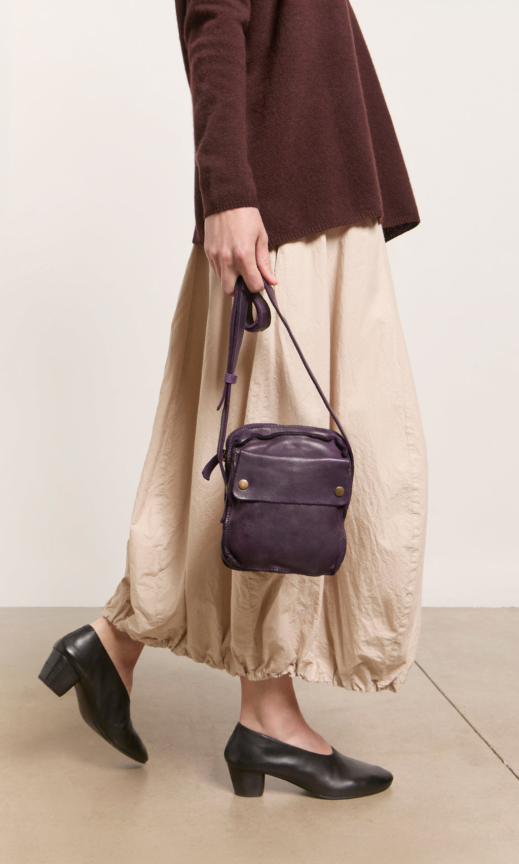 Plümo  Person in a brown sweater, beige skirt, holding the Loyd Bag, and wearing black heeled shoes.