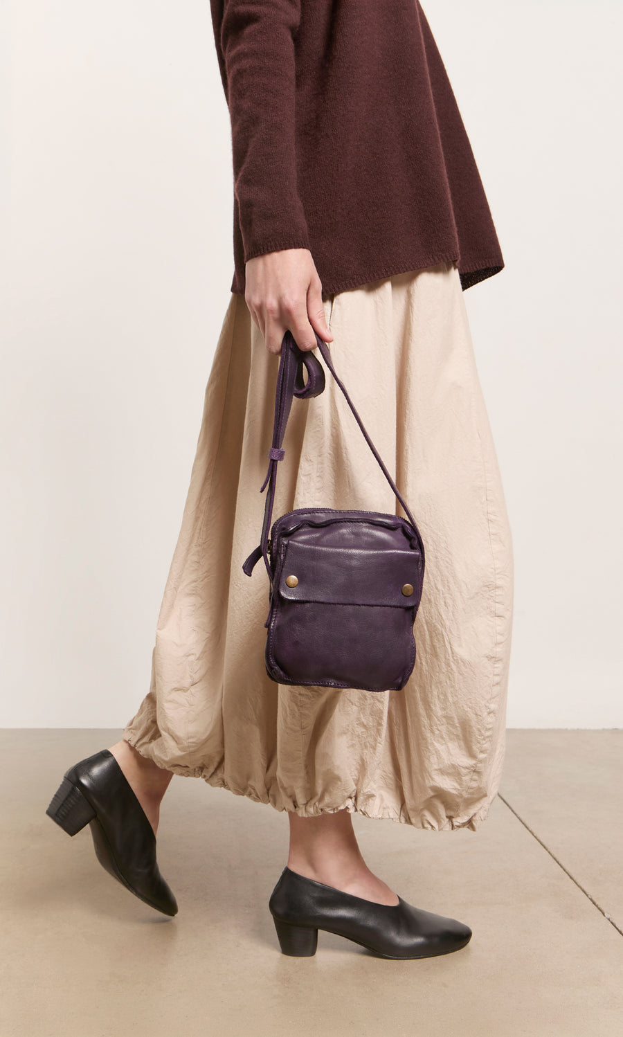 Plümo  Person in a brown sweater, beige skirt, holding the Loyd Bag, and wearing black heeled shoes.