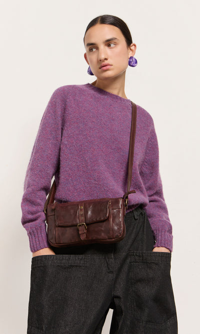Plümo  Person in a purple sweater, black pants, purple earrings, and a Luxton Bag, hands in pockets.