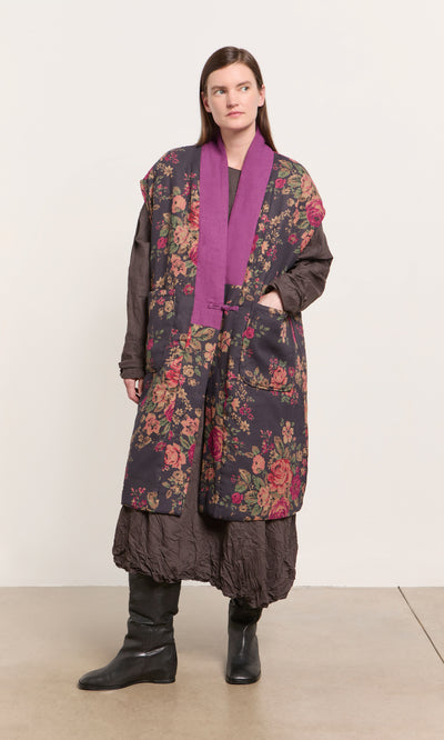 Plümo  Woman in a Manton Gilet with pink trim, brown dress, and black boots stands by a plain light wall.