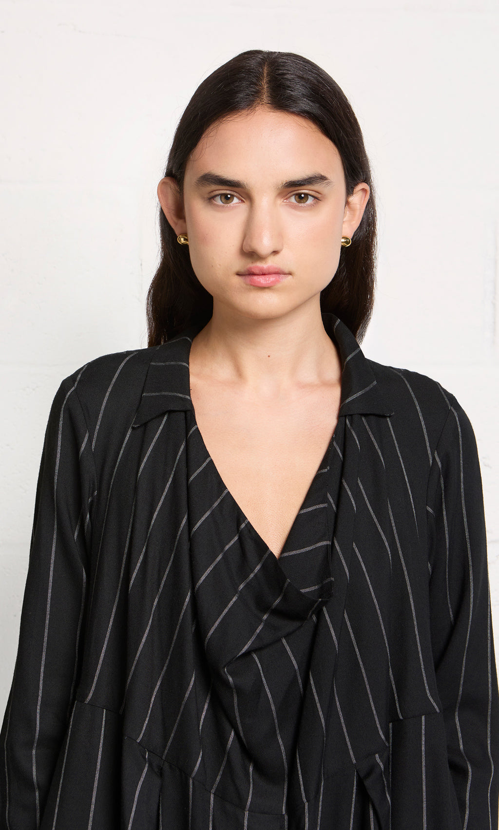 Plümo  Woman with long dark hair wears the Manza Dress, inspired by menswear suiting, standing against a white wall.