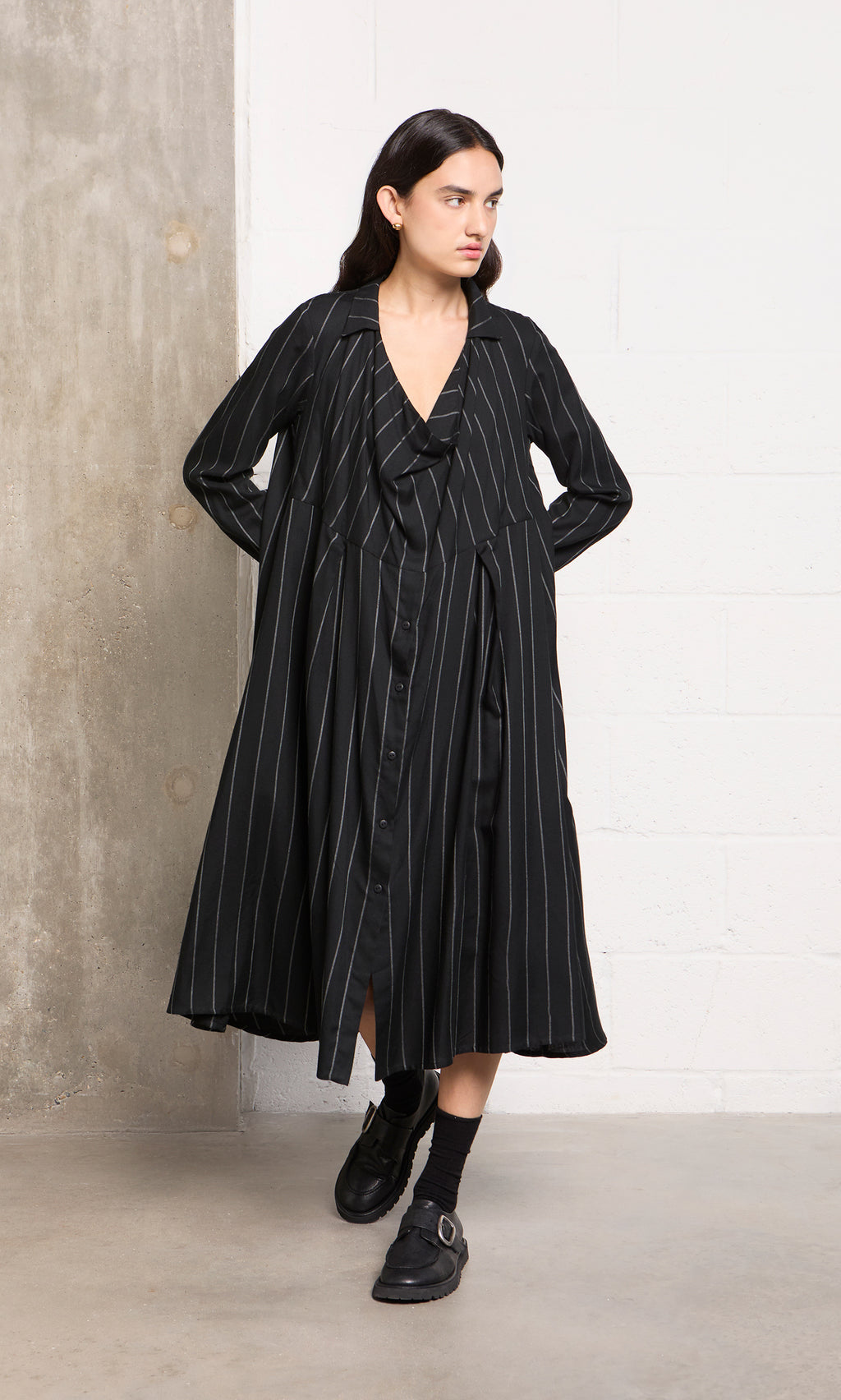 Plümo  Woman wears the Manza Dress, a black pinstriped button-front tunic with menswear vibes, by a concrete wall.
