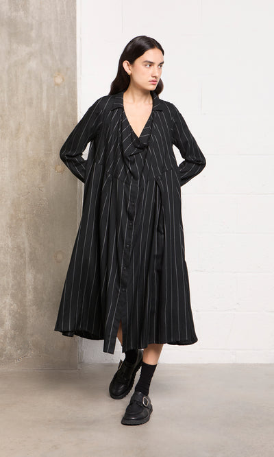 Plümo  Woman wears the Manza Dress, a black pinstriped button-front tunic with menswear vibes, by a concrete wall.