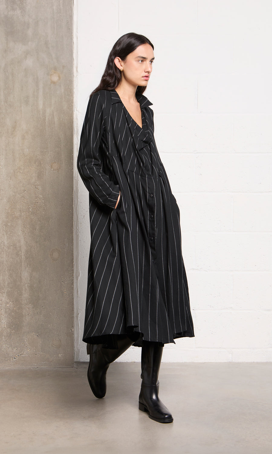 Plümo  Person in the Manza Dress and black boots stands by a concrete and white brick wall.