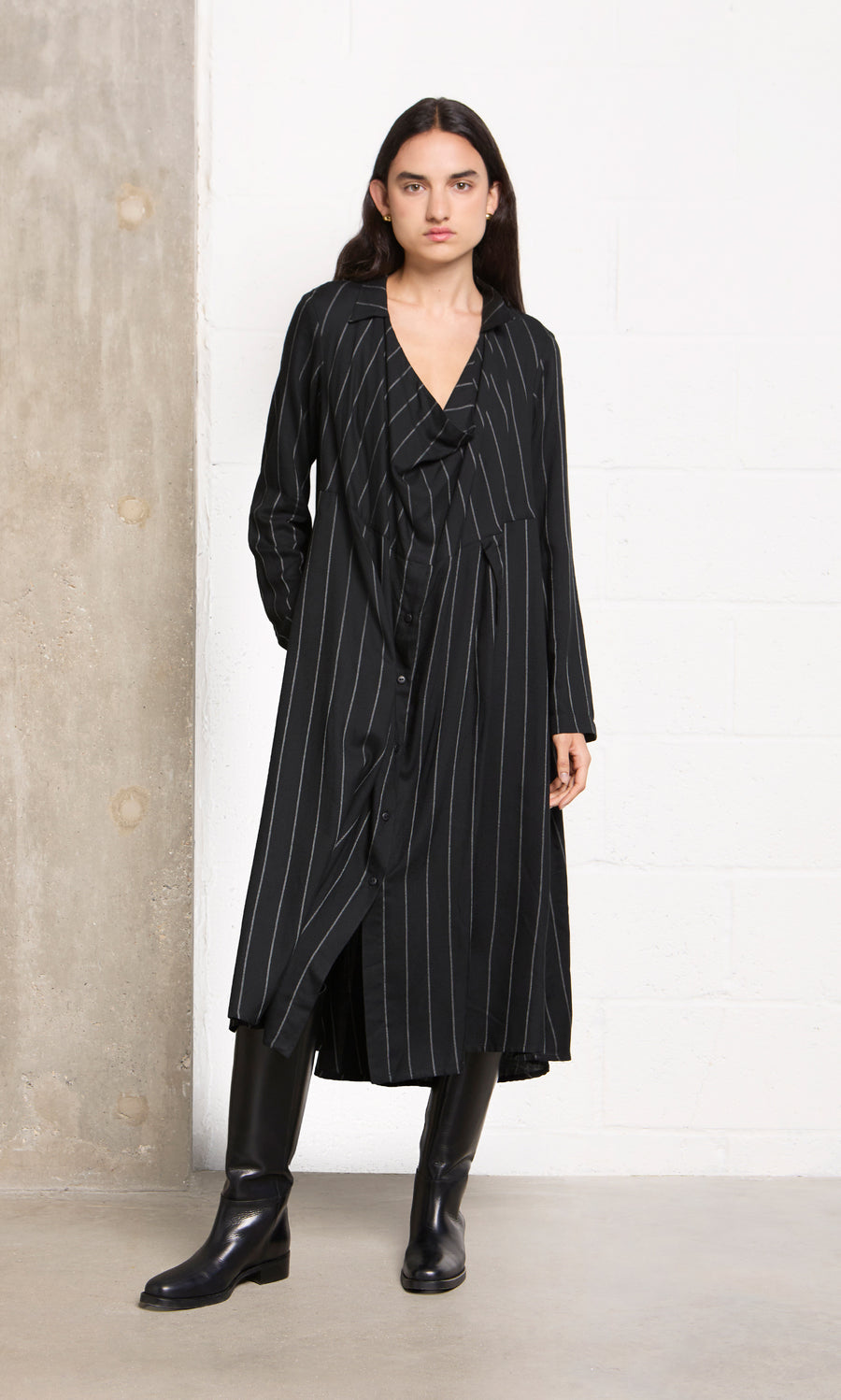 Plümo  Person in the Manza Dress and black boots stands against a white wall and concrete pillar.