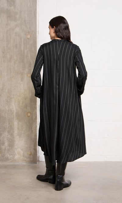 Plümo  Someone in the Manza Dress and black boots faces a concrete wall, channeling classic menswear-inspired style.