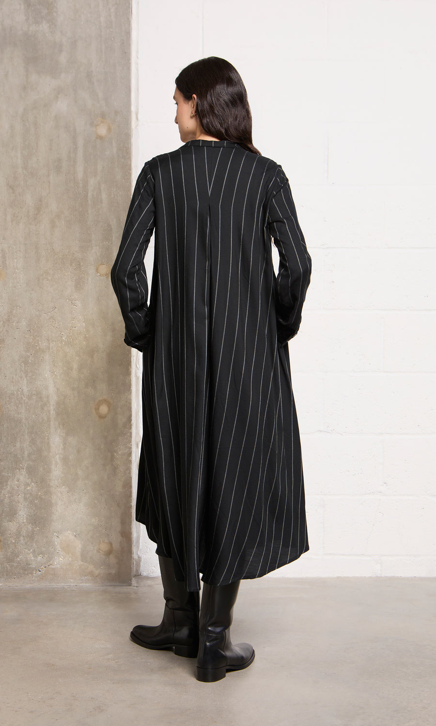 Plümo  Someone in the Manza Dress and black boots faces a concrete wall, channeling classic menswear-inspired style.