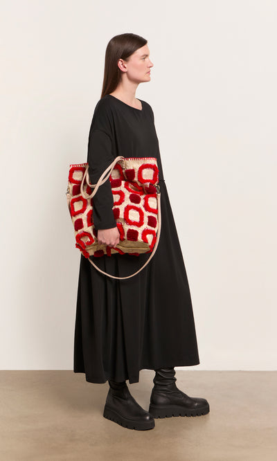 Plümo  Woman in a long black dress holds the Marakech Bag, an embroidered tote made in Morocco, against a plain background.