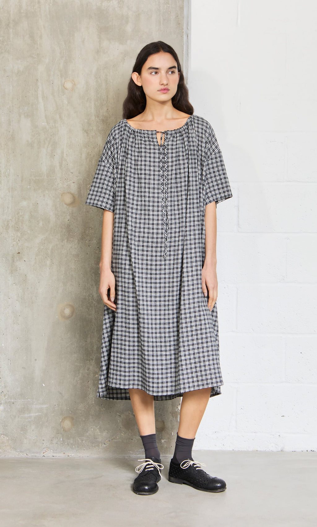 Plümo- Woman wears the Marin Dress - Plaid, a loose black-and-white short-sleeve dress made in London, with black shoes.