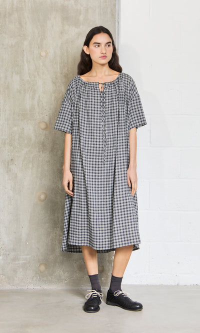 Plümo- Woman wears the Marin Dress - Plaid, a loose black-and-white short-sleeve dress made in London, with black shoes.