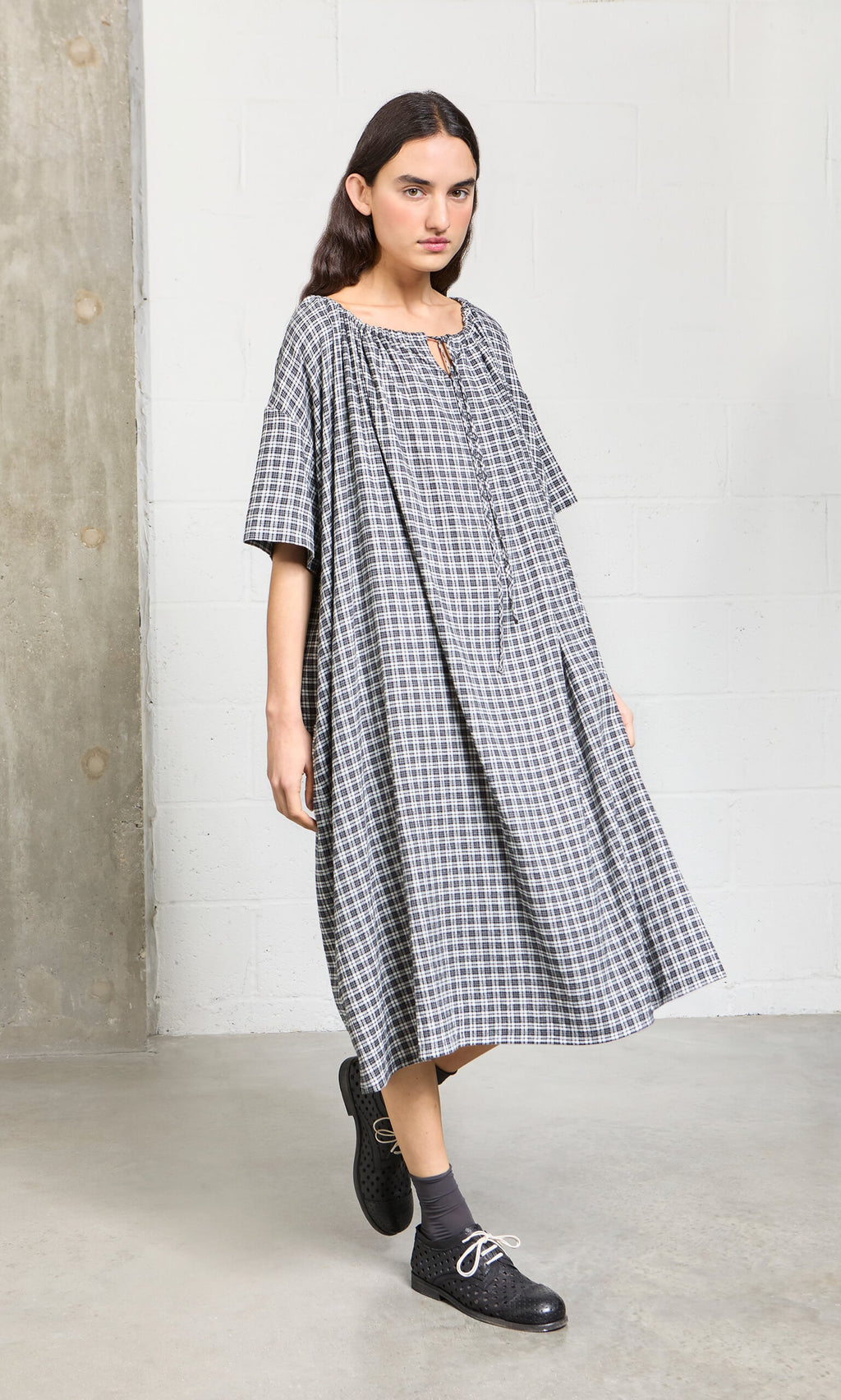 Plümo- Woman in the Marin Dress - Plaid, with black shoes and gray socks, standing indoors by a white wall and concrete pillar.
