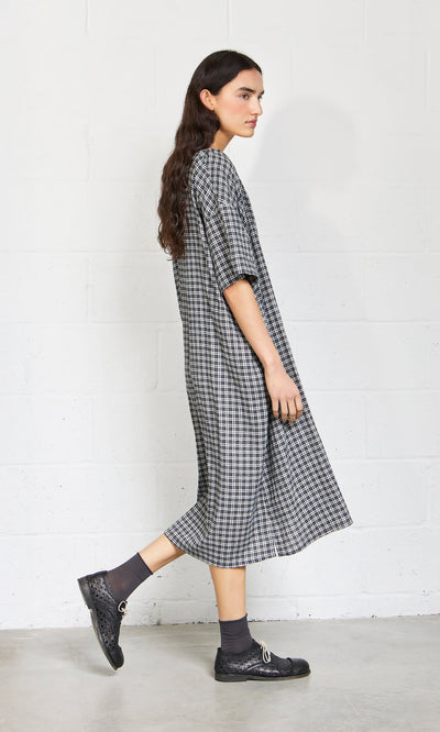 Plümo- A woman in the Marin Dress - Plaid stands sideways by a white brick wall, wearing black shoes and gray socks.