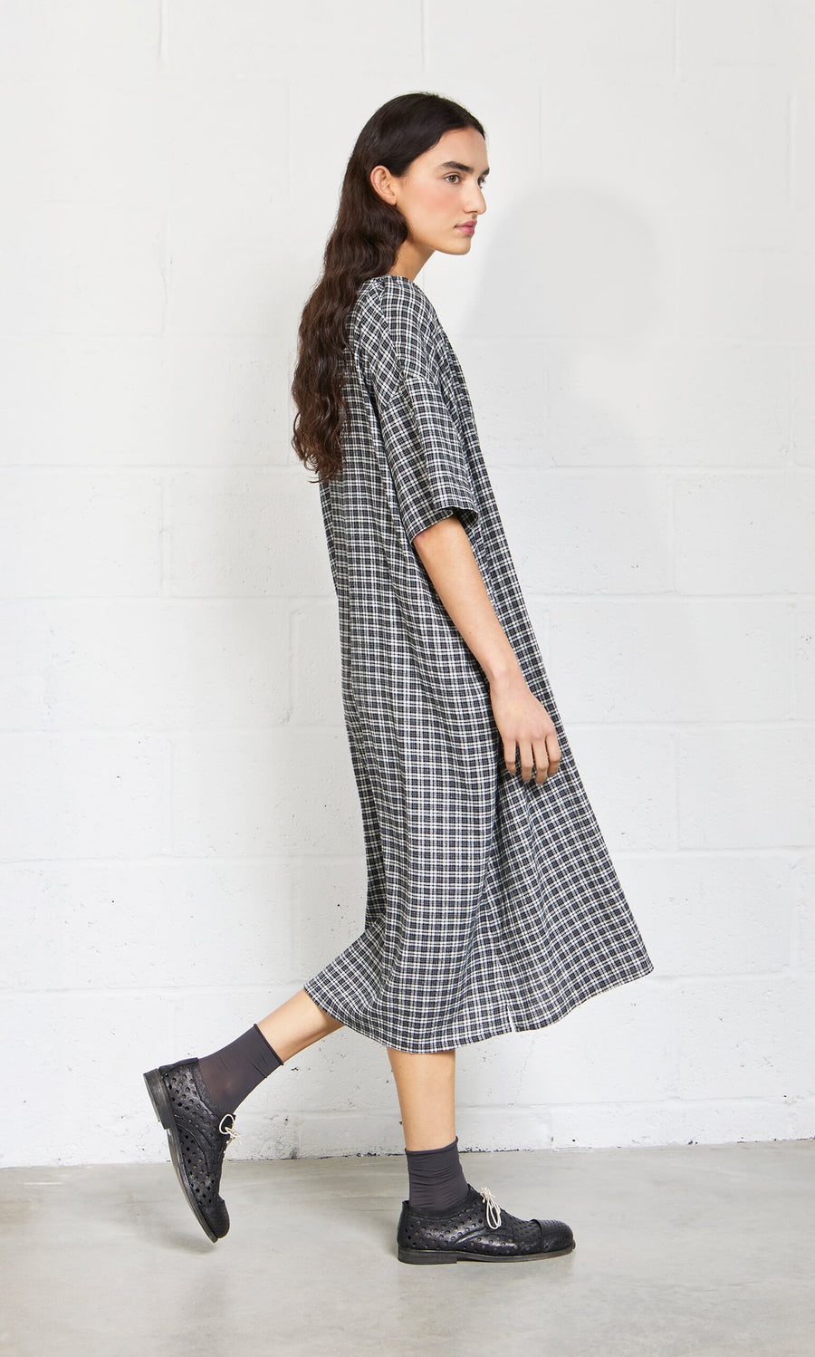 Plümo- A woman in the Marin Dress - Plaid stands sideways by a white brick wall, wearing black shoes and gray socks.