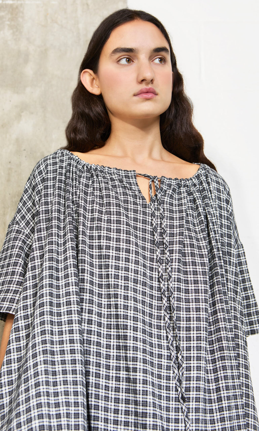 Plümo- Woman with long dark hair wears the Marin Dress - Plaid, a sustainable hemp piece made in London, against a pale backdrop.
