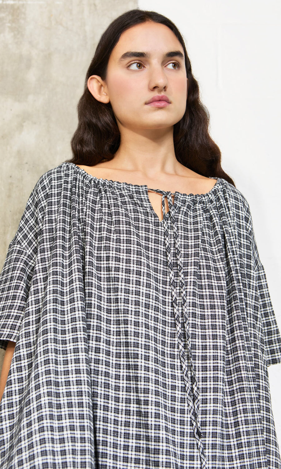 Plümo- Woman with long dark hair wears the Marin Dress - Plaid, a sustainable hemp piece made in London, against a pale backdrop.