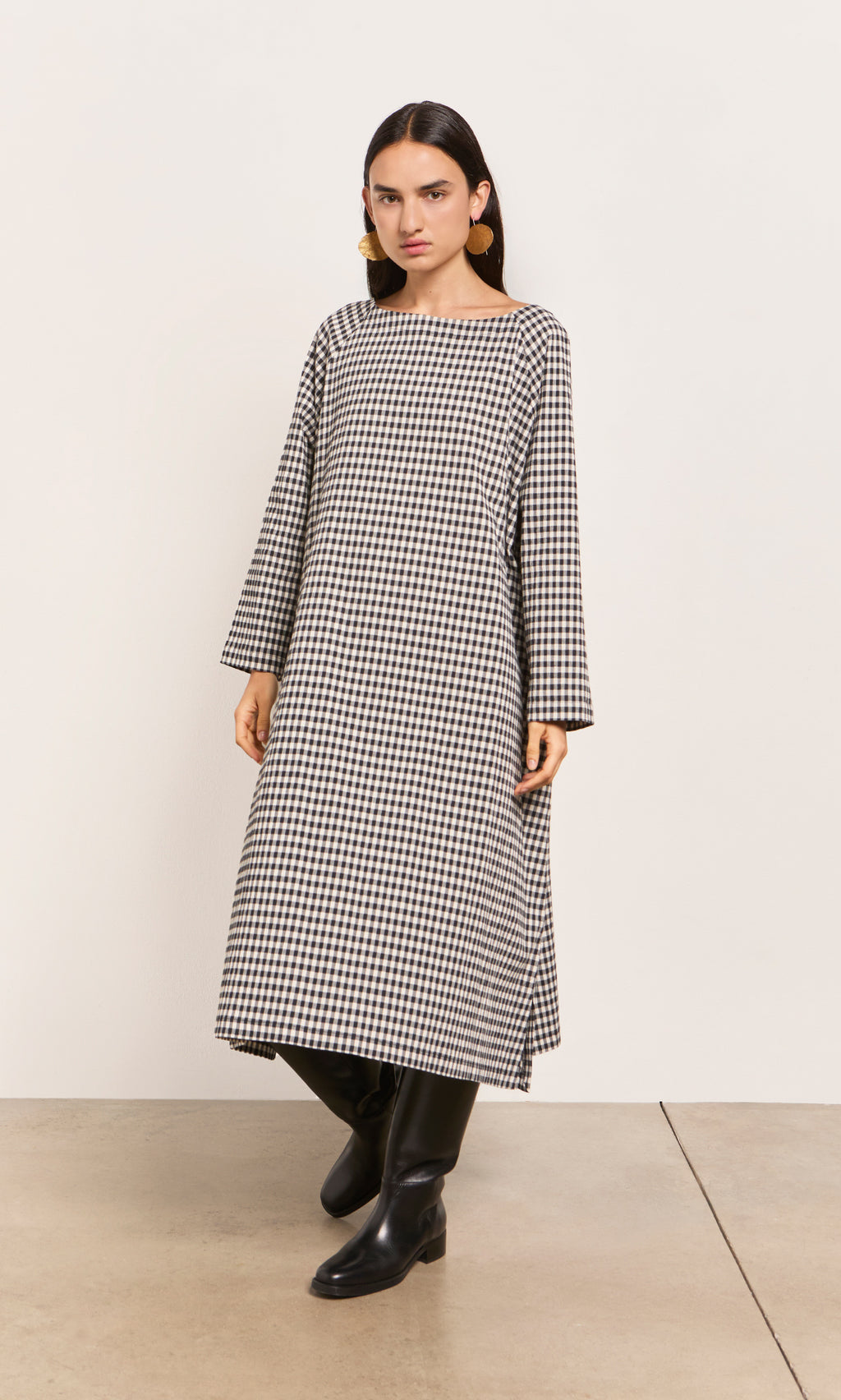 Plümo  Woman in Marsell Dress - Gingham, loose fit, slash neck, with black boots indoors against a plain wall.